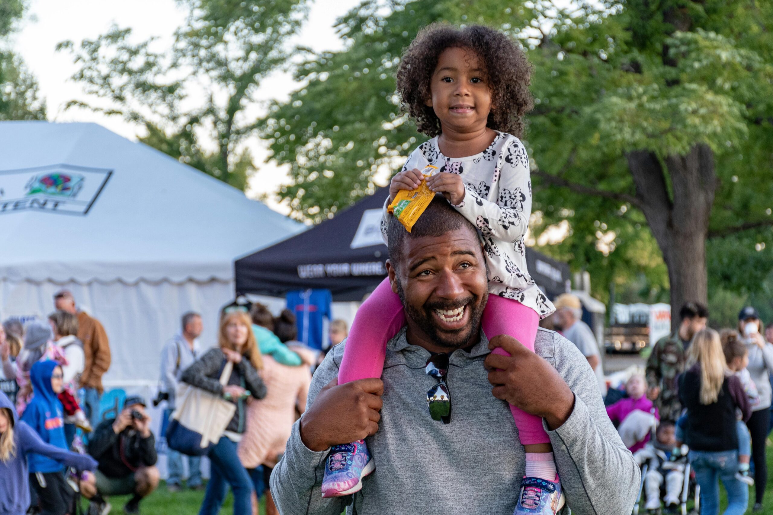 A joyful father carrying his daughter at an outdoor festival surrounded by people and tents.